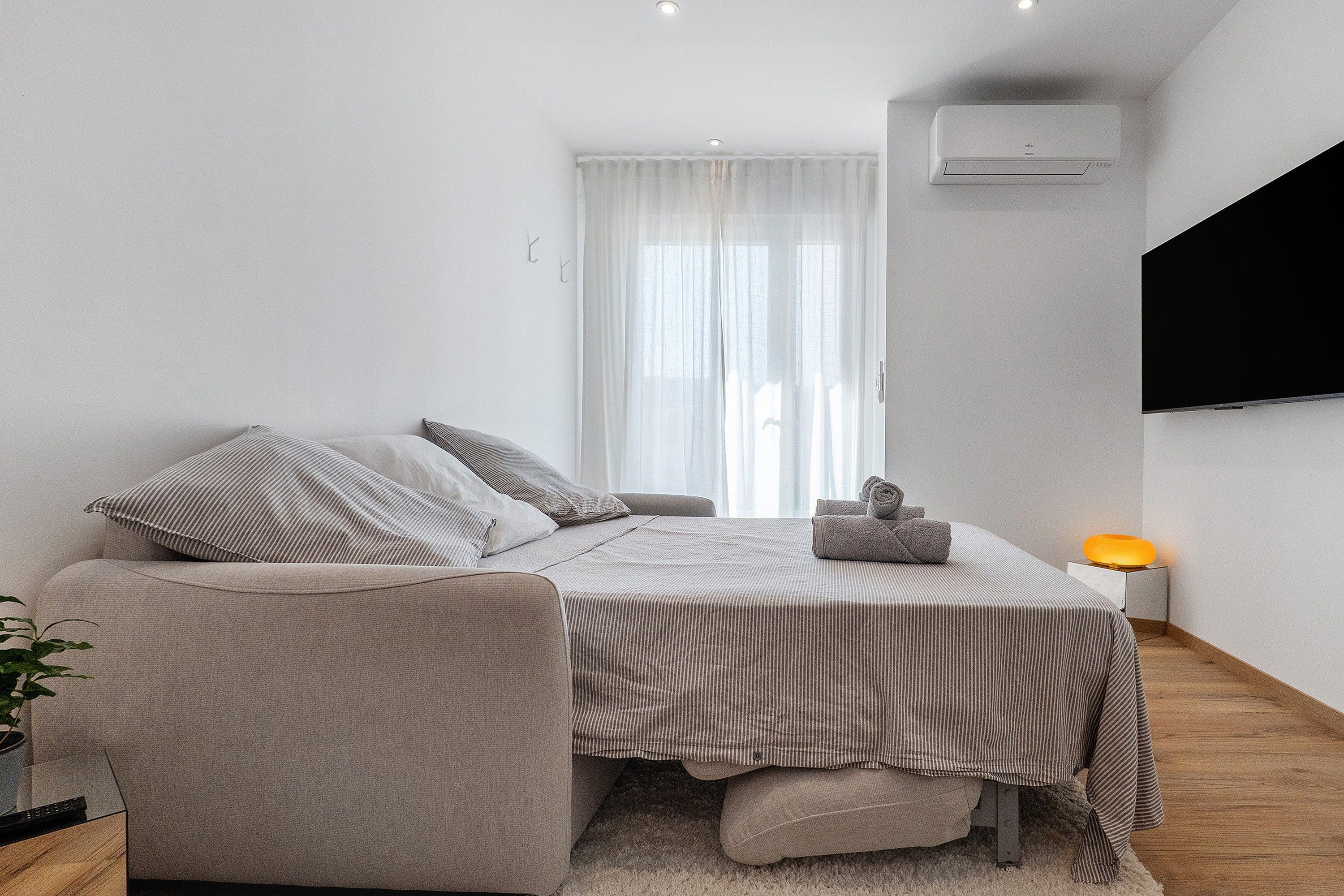 Une chambre à coucher moderne, avec un lit, une chaise, une table de chevet avec une lampe et une grande fenêtre avec des rideaux.