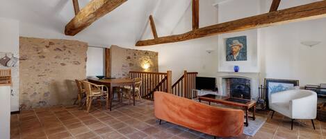 A rustic interior with exposed wooden beams, a stone wall, a fireplace, and a dining area with wicker furniture.