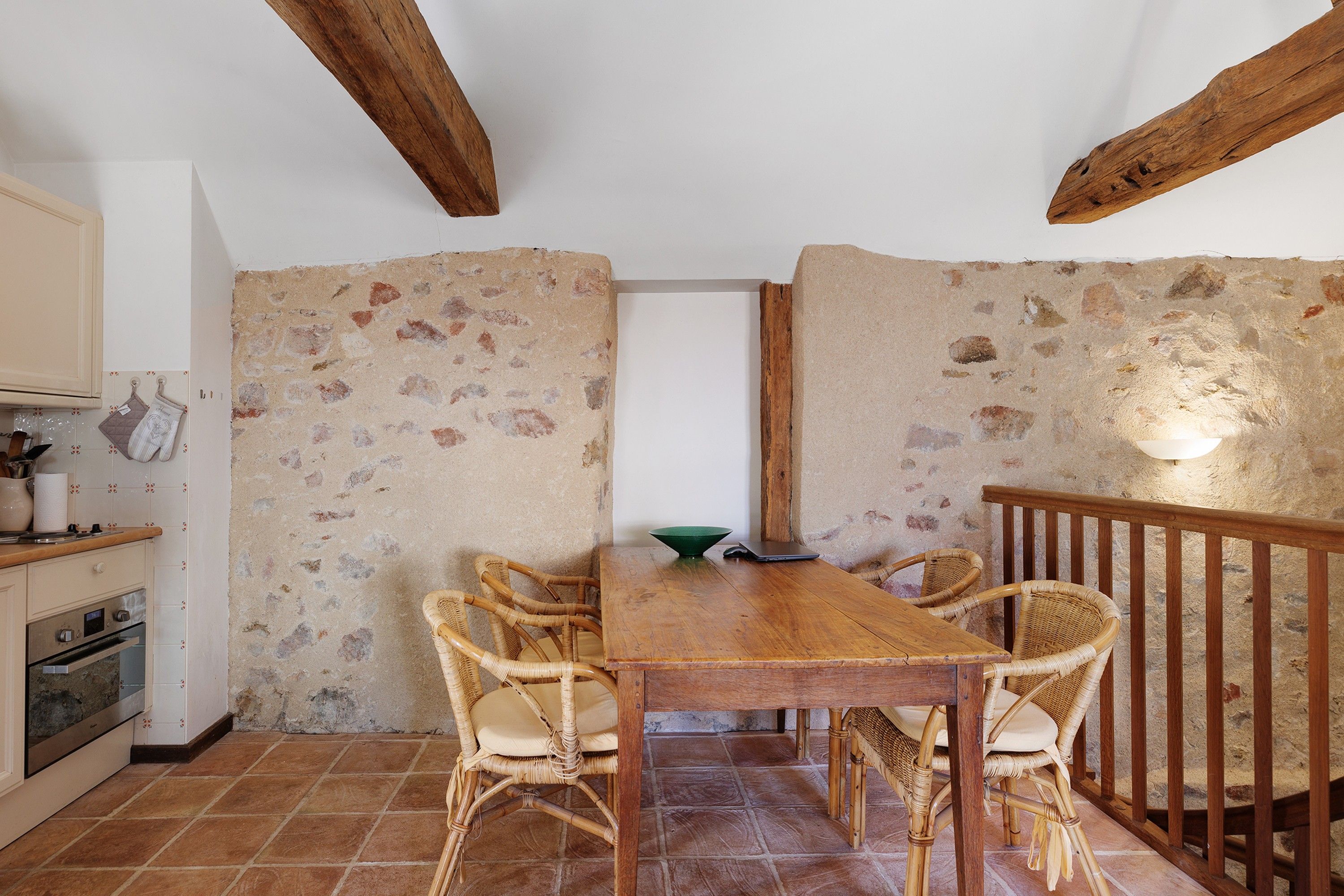 A rustic kitchen with a wooden table, wicker chairs, and a stone wall.