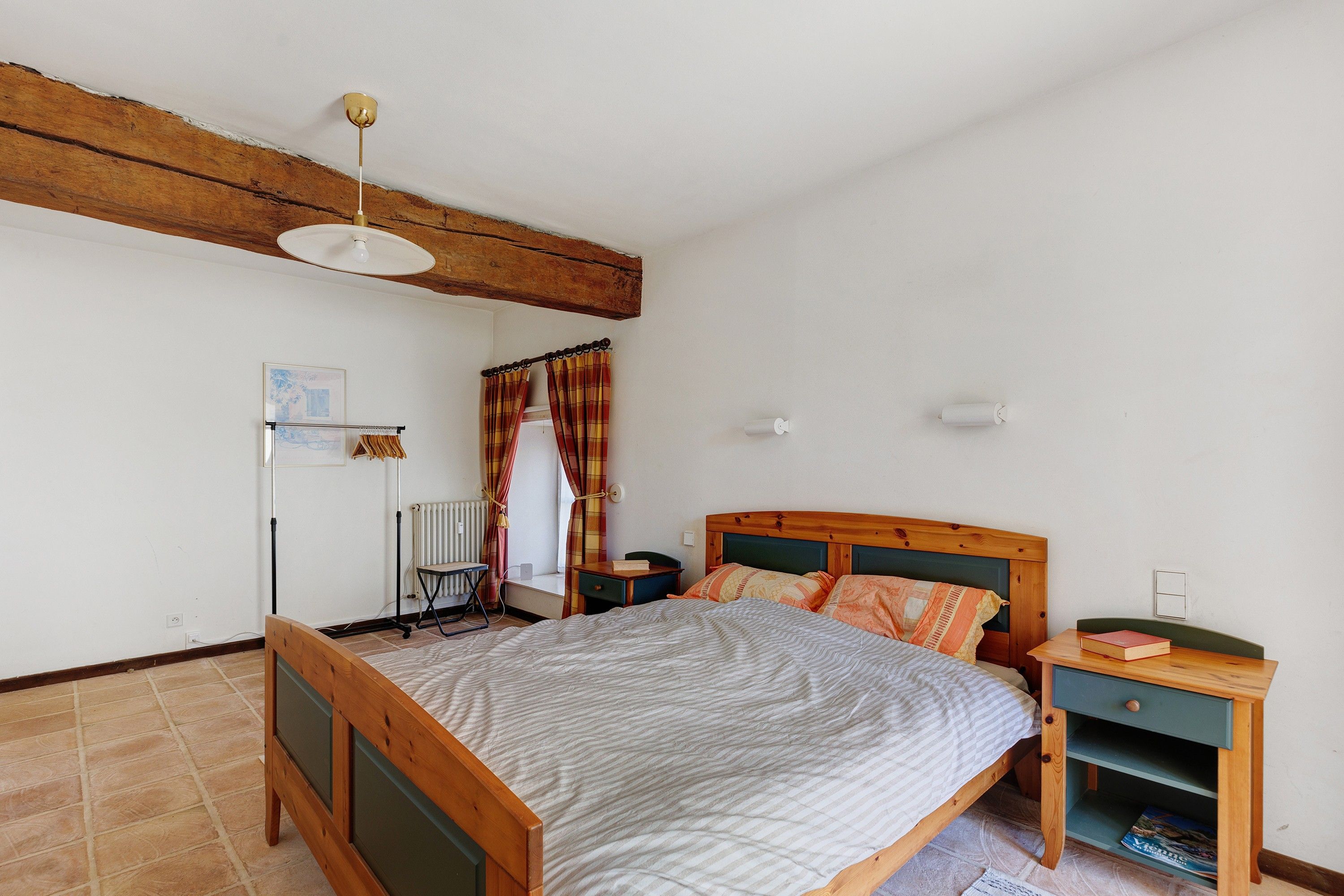 Une chambre à coucher avec un lit en bois, une table de chevet, une commode, un radiateur, une fenêtre avec des rideaux et un ventilateur de plafond.