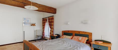 A bedroom with a wooden bed, a bedside table, a nightstand, a radiator, a window with curtains, and a ceiling fan.