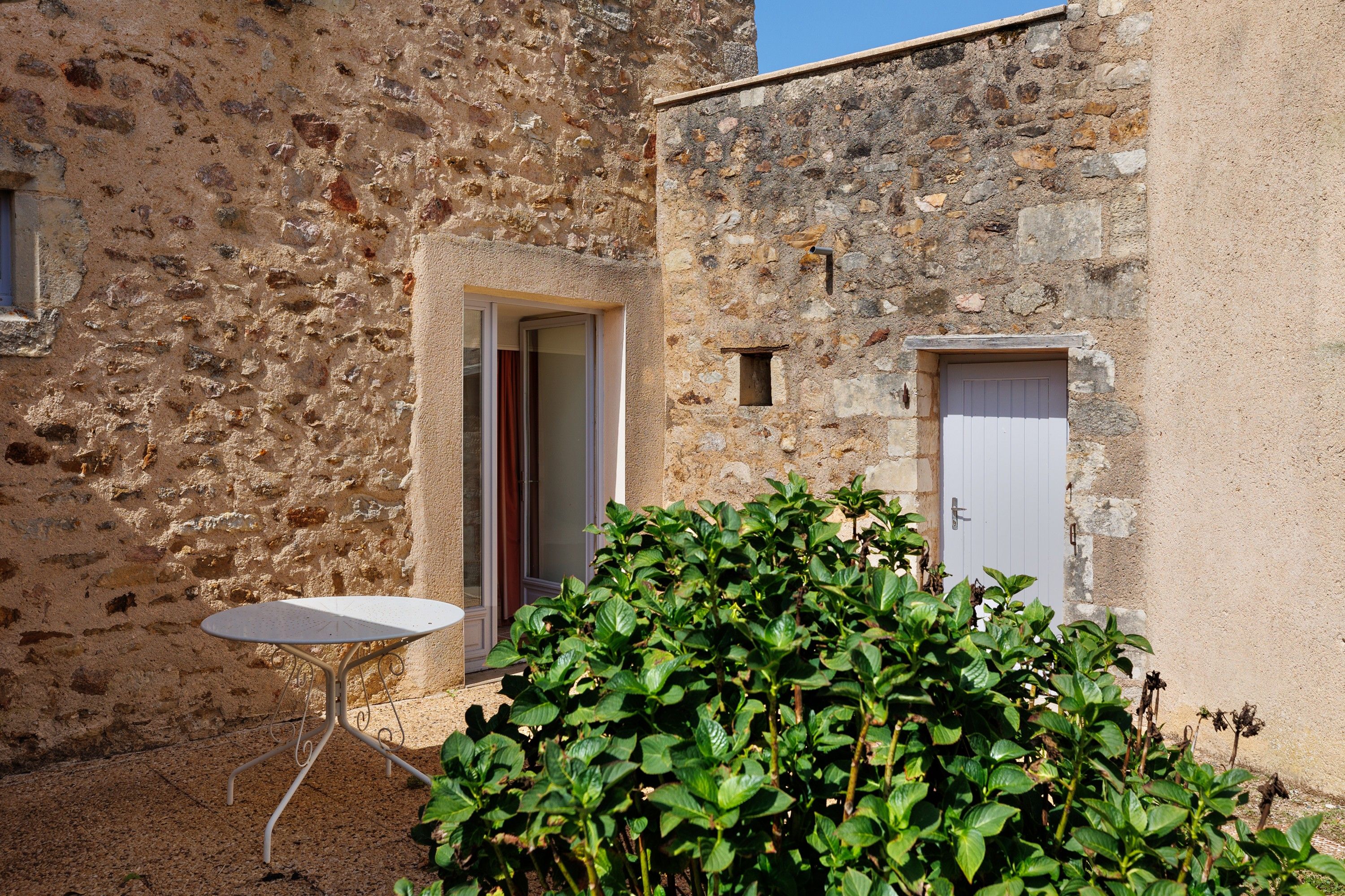 Un edificio de piedra con una puerta y una ventana, una mesa pequeña, y un arbusto al frente.