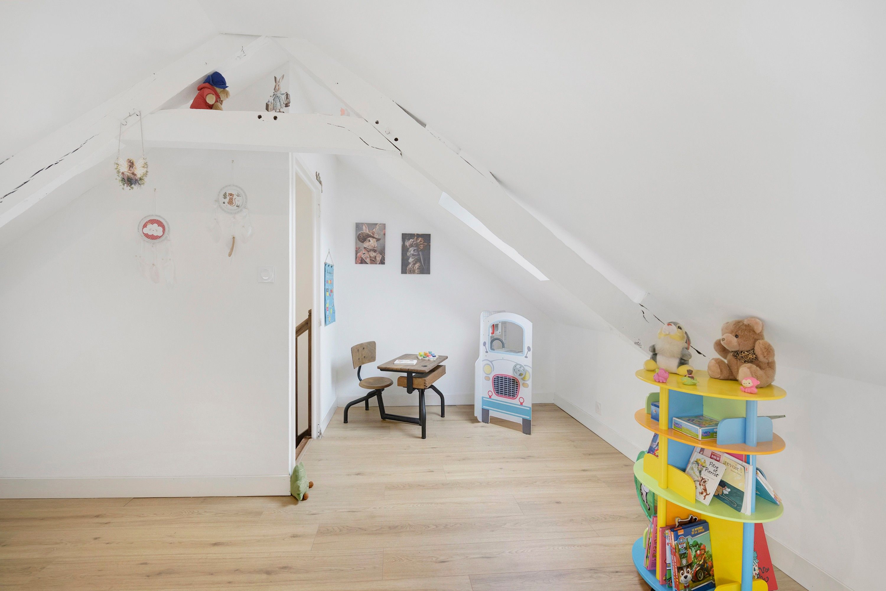 Una habitación con techo inclinado, suelo de madera, una pequeña mesa con una silla y una estructura de juguete colorida.