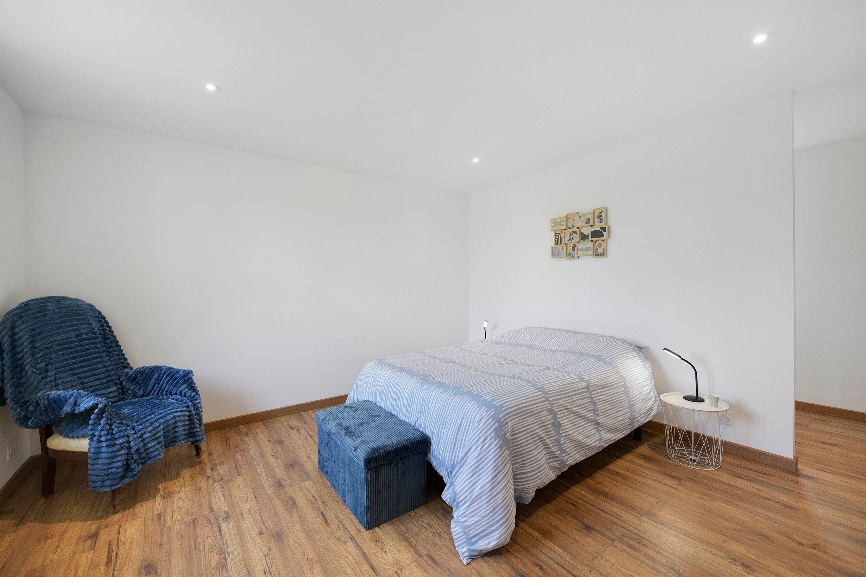 Een slaapkamer met een bed, een stoel, een klein tafeltje met een lamp en een kunstwerk aan de muur.
