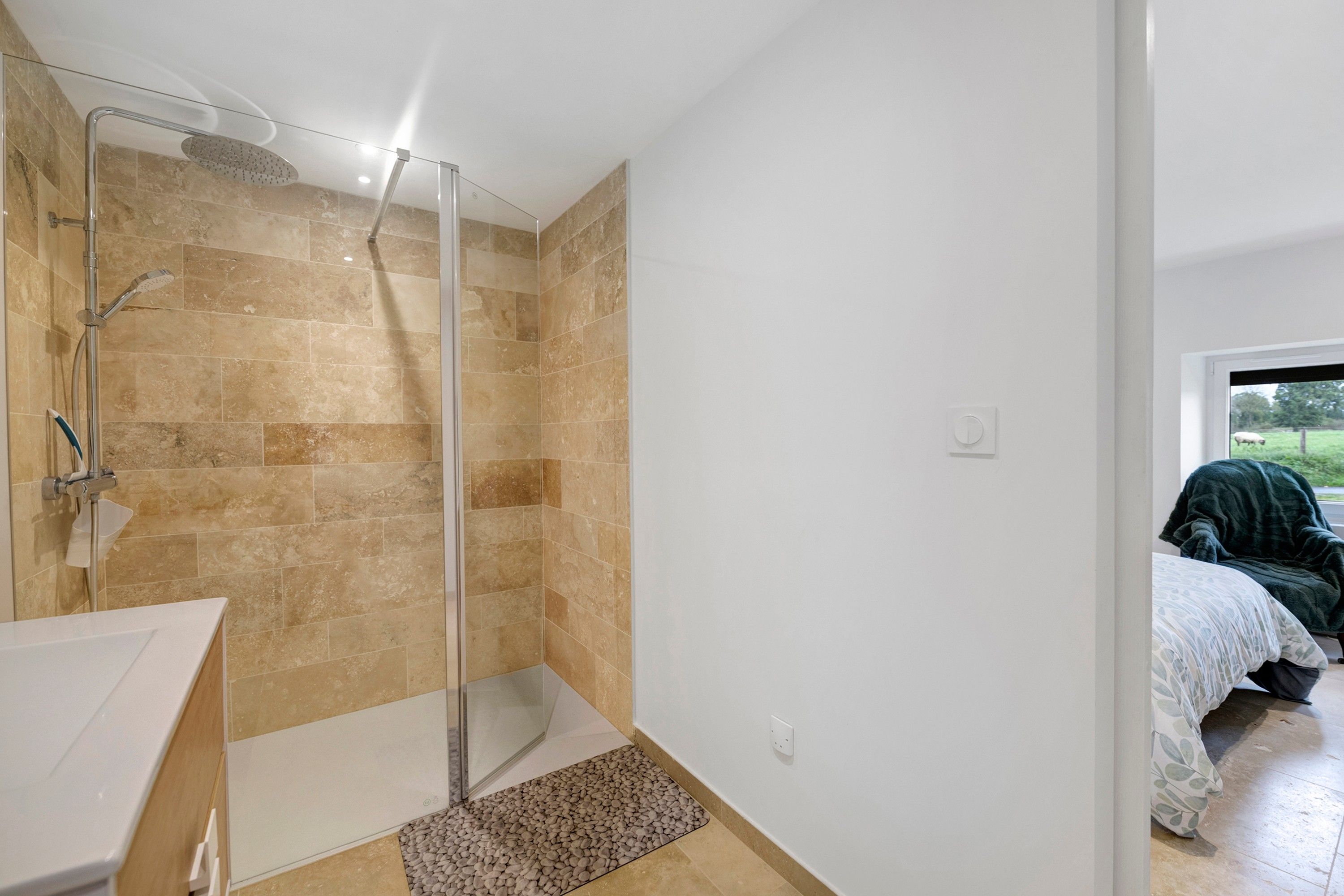Une salle de bain avec une douche, un lavabo et une fenêtre donnant sur une chambre.