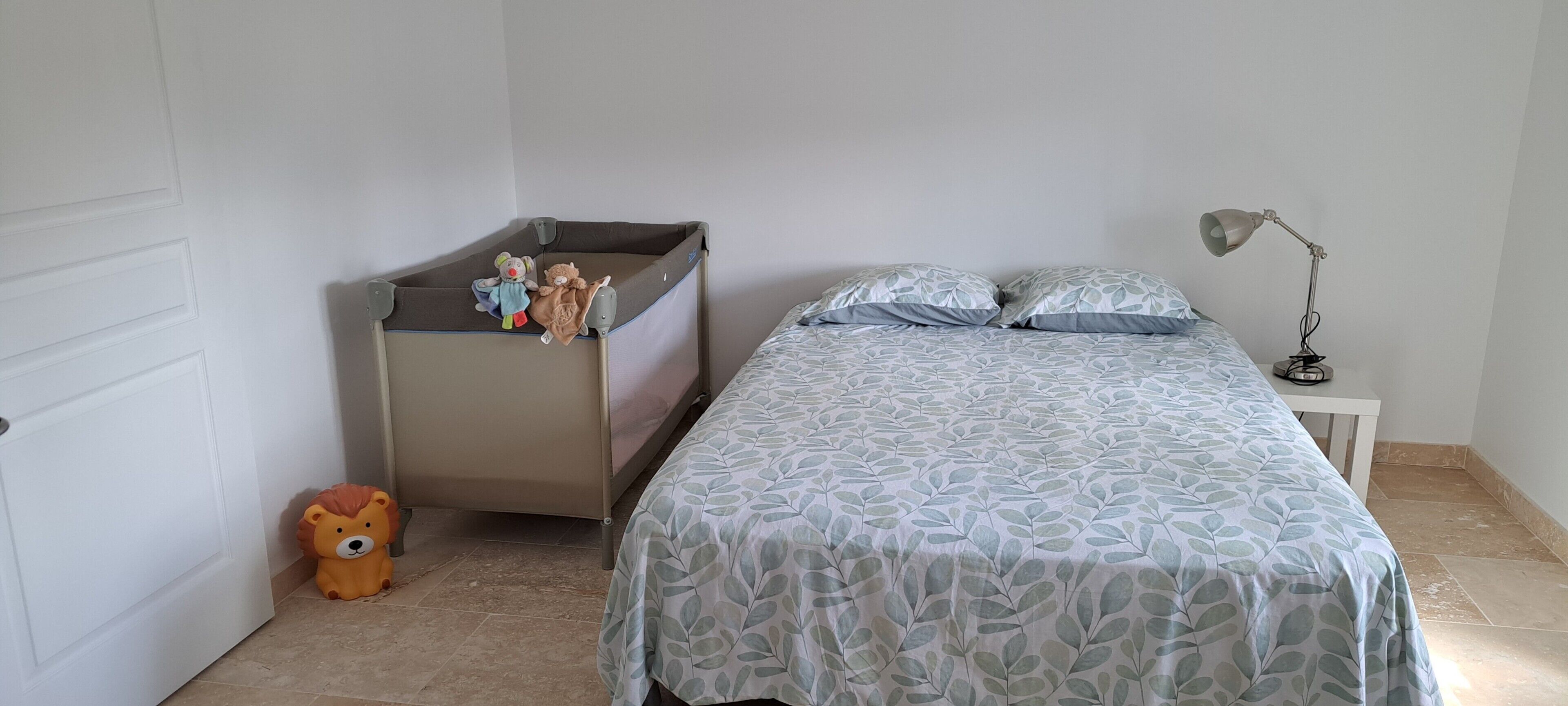 A bedroom with a bed, a nightstand, a lamp, a crib, and a teddy bear.
