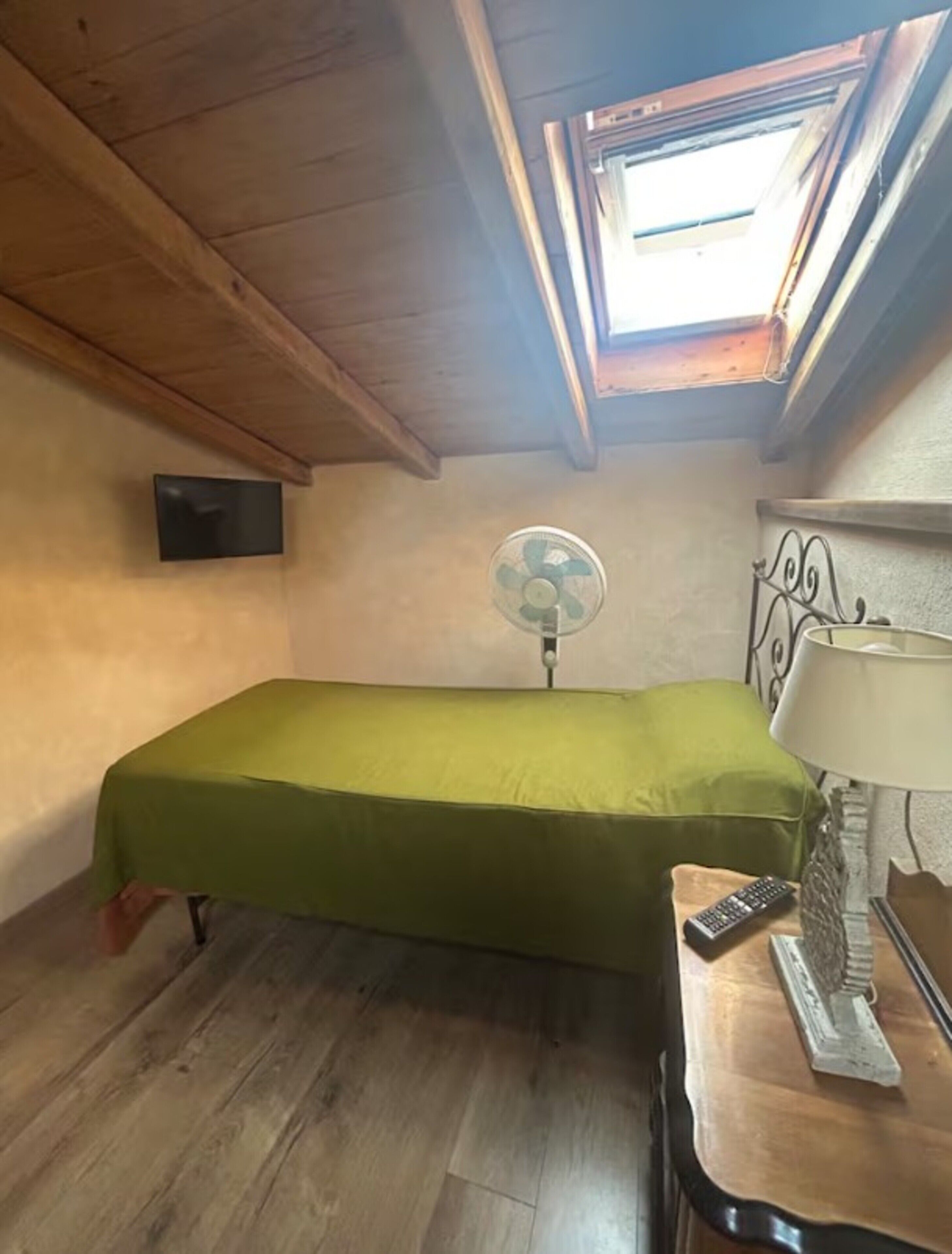 Une petite chambre avec un lit simple, un plafond en bois, un puits de lumière, un téléviseur fixé au mur, un ventilateur sur pied et une lampe de chevet.