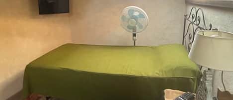 Une petite chambre avec un lit simple, un plafond en bois, un puits de lumière, un téléviseur fixé au mur, un ventilateur sur pied et une lampe de chevet.