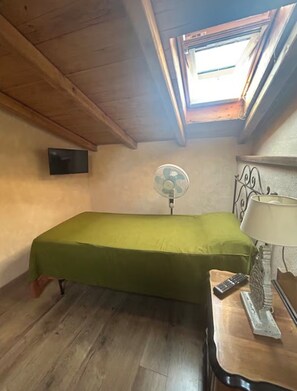 Ein kleines Zimmer mit einem Einzelbett, Holzboden, einem Oberlicht, einem Wand-Fernseher, einem Standventilator und einer Nachttischlampe.