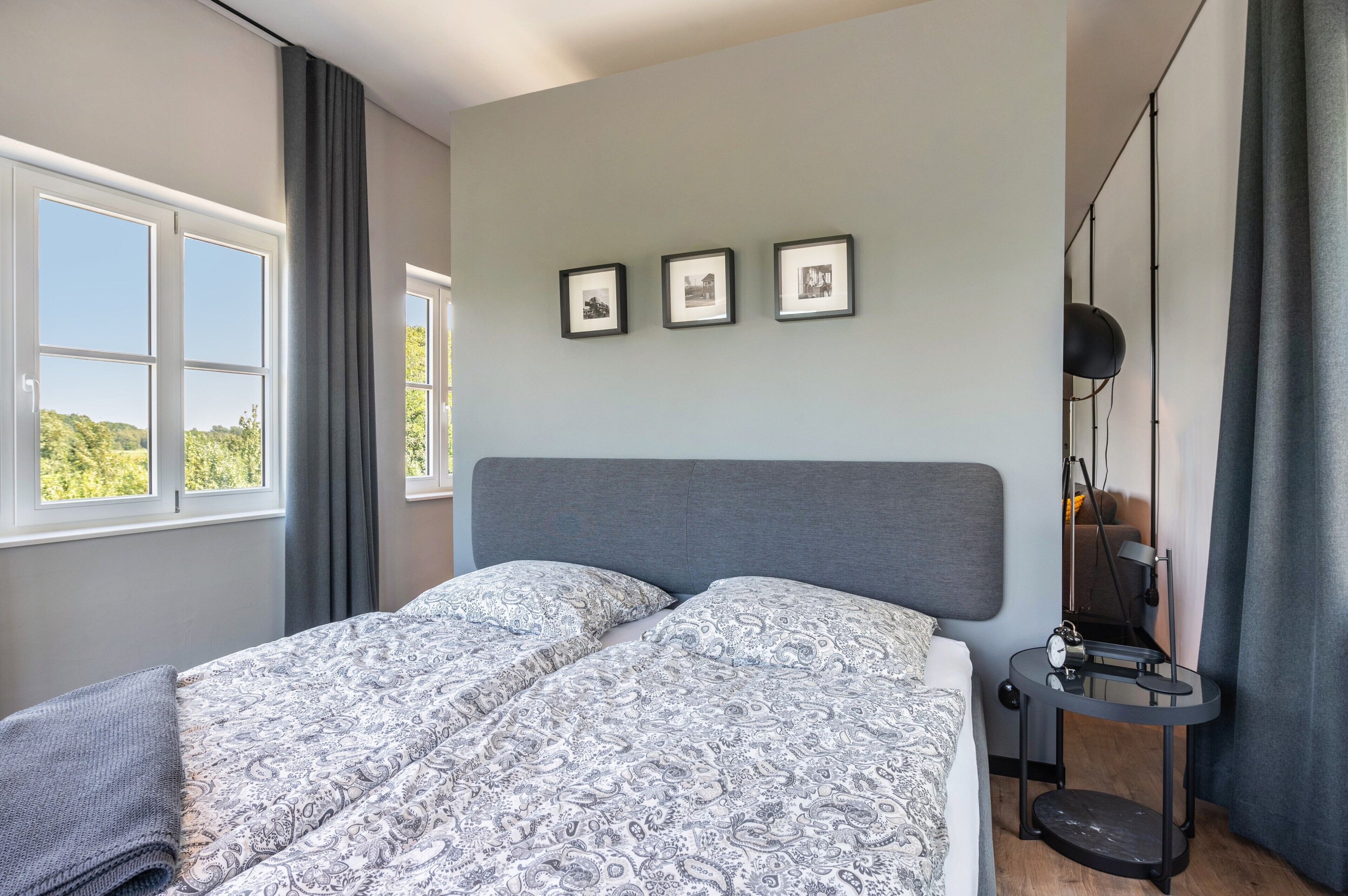 Een slaapkamer met een bed, twee nachtkastjes, een vloerlamp en drie foto’s in lijsten aan de muur.