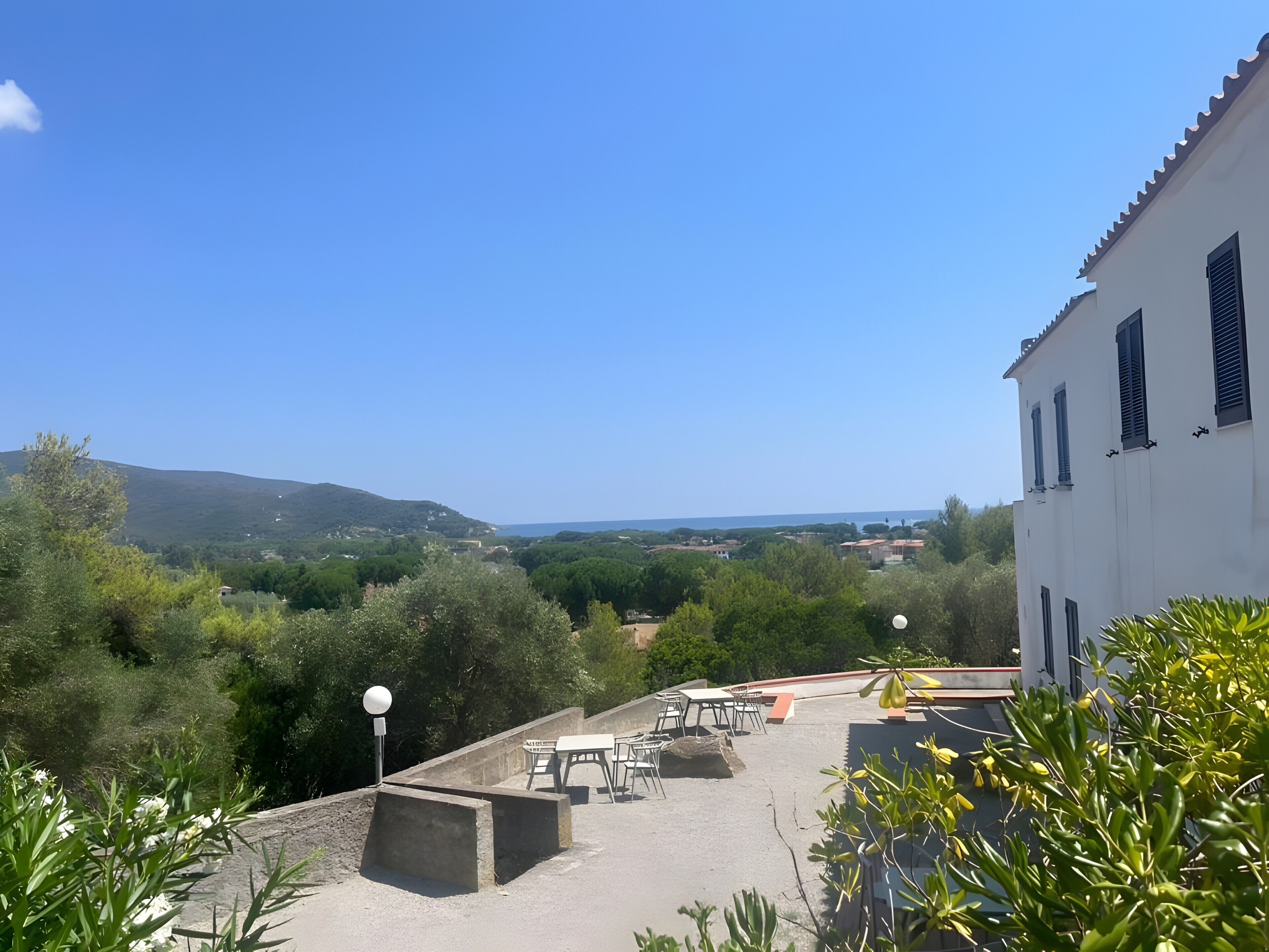 Una terraza con mobiliario exterior, un edificio blanco con persianas y vistas a un paisaje montañoso.