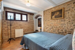 Un dormitorio con pared de piedra, una cama con cubrecama azul, un radiador y una ventana con vistas.