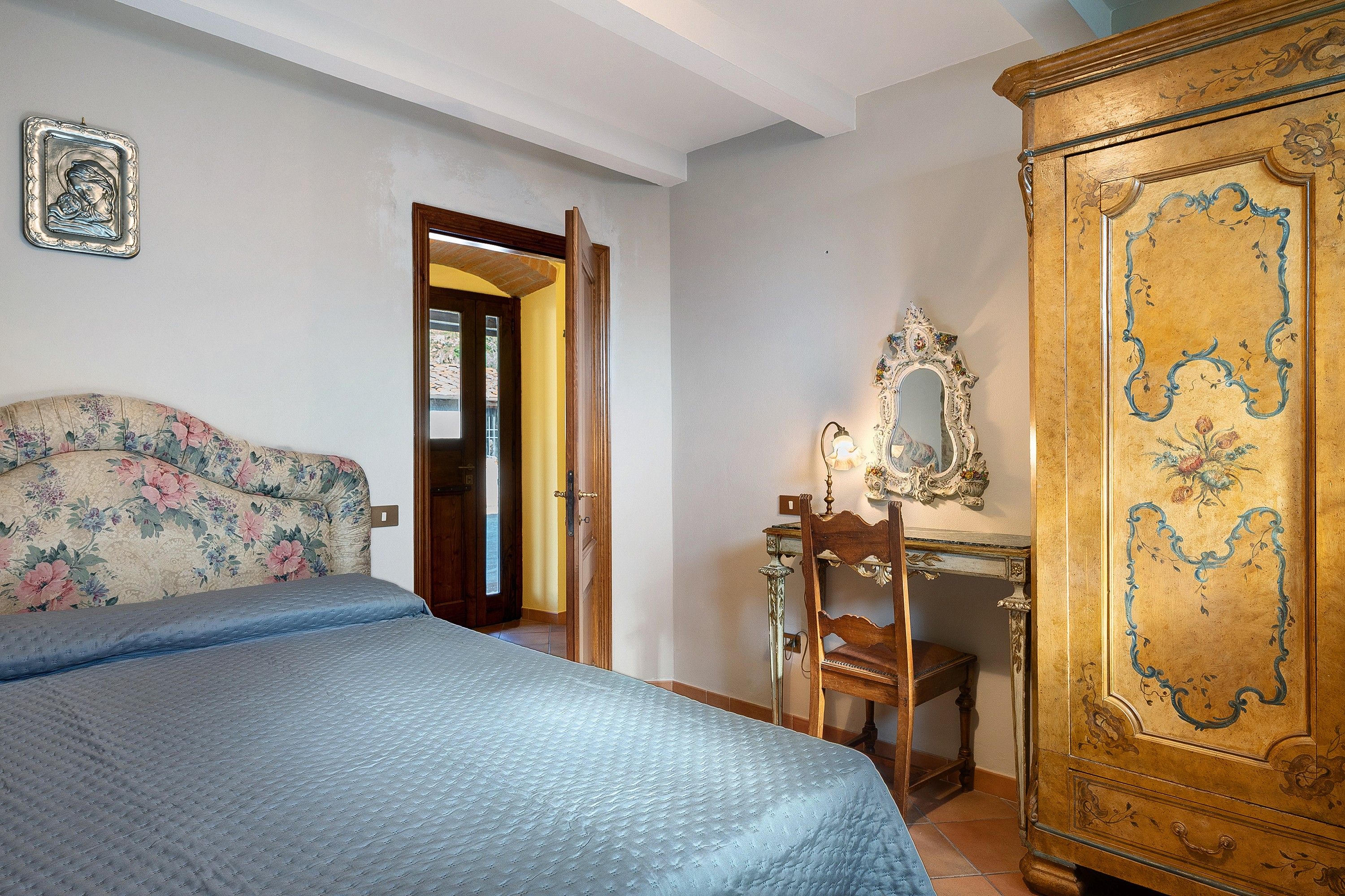 Een slaapkamer met een bed, een houten garderobe, een bureau met spiegel en een deur naar een andere kamer.