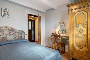 A bedroom with a bed, a wooden wardrobe, a desk with a mirror, and a door leading to another room.