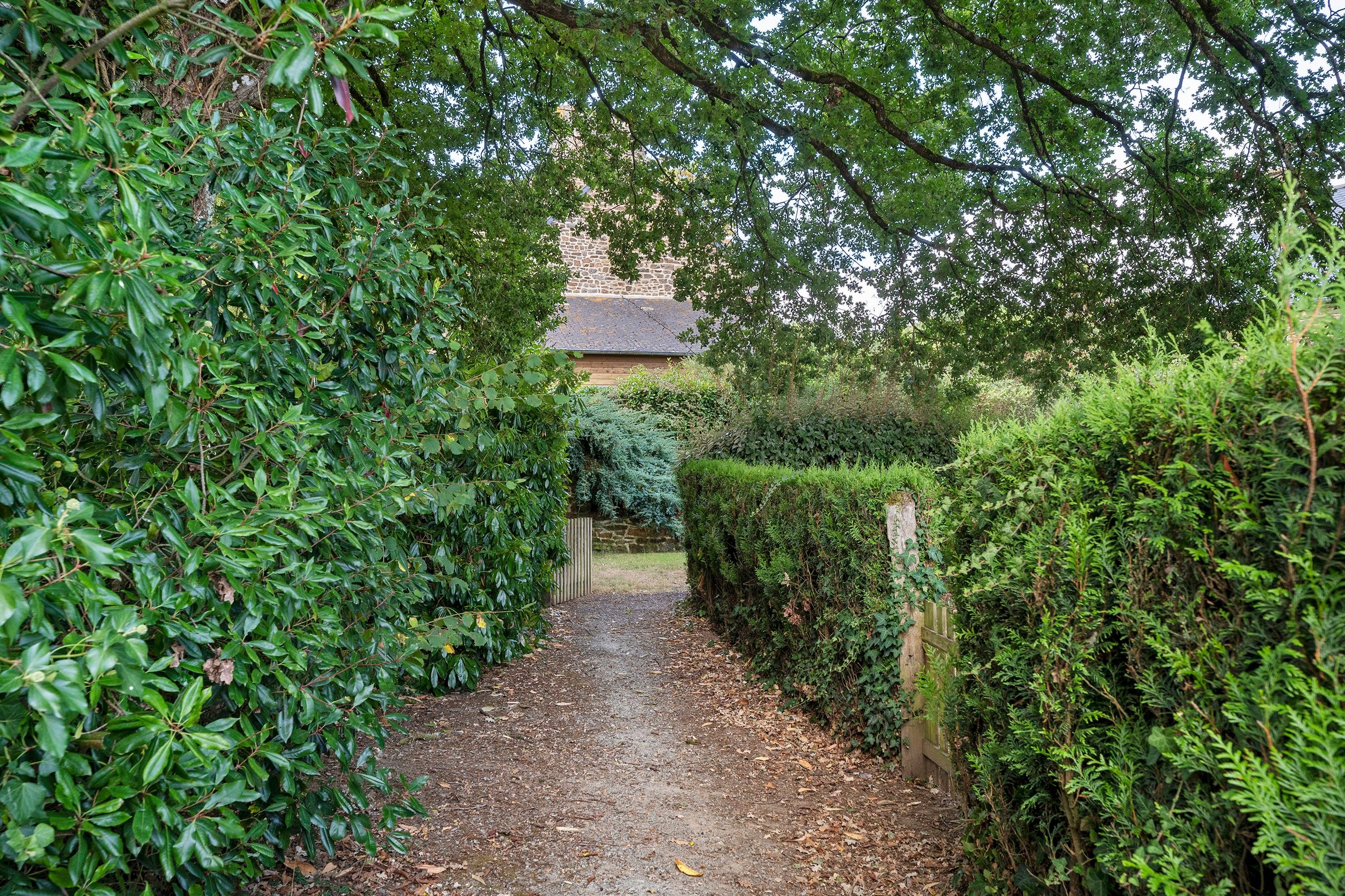 Un sentier étroit, bordé d’une végétation luxuriante, mène à un bâtiment doté d’un mur en pierre et d’un toit à deux versants.