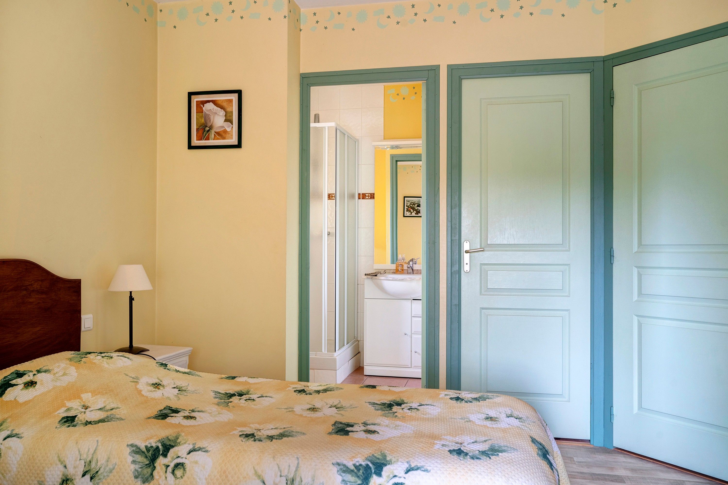 Quarto com cama, mesa de cabeceira, luminária, porta e banheiro visível através de uma porta aberta.