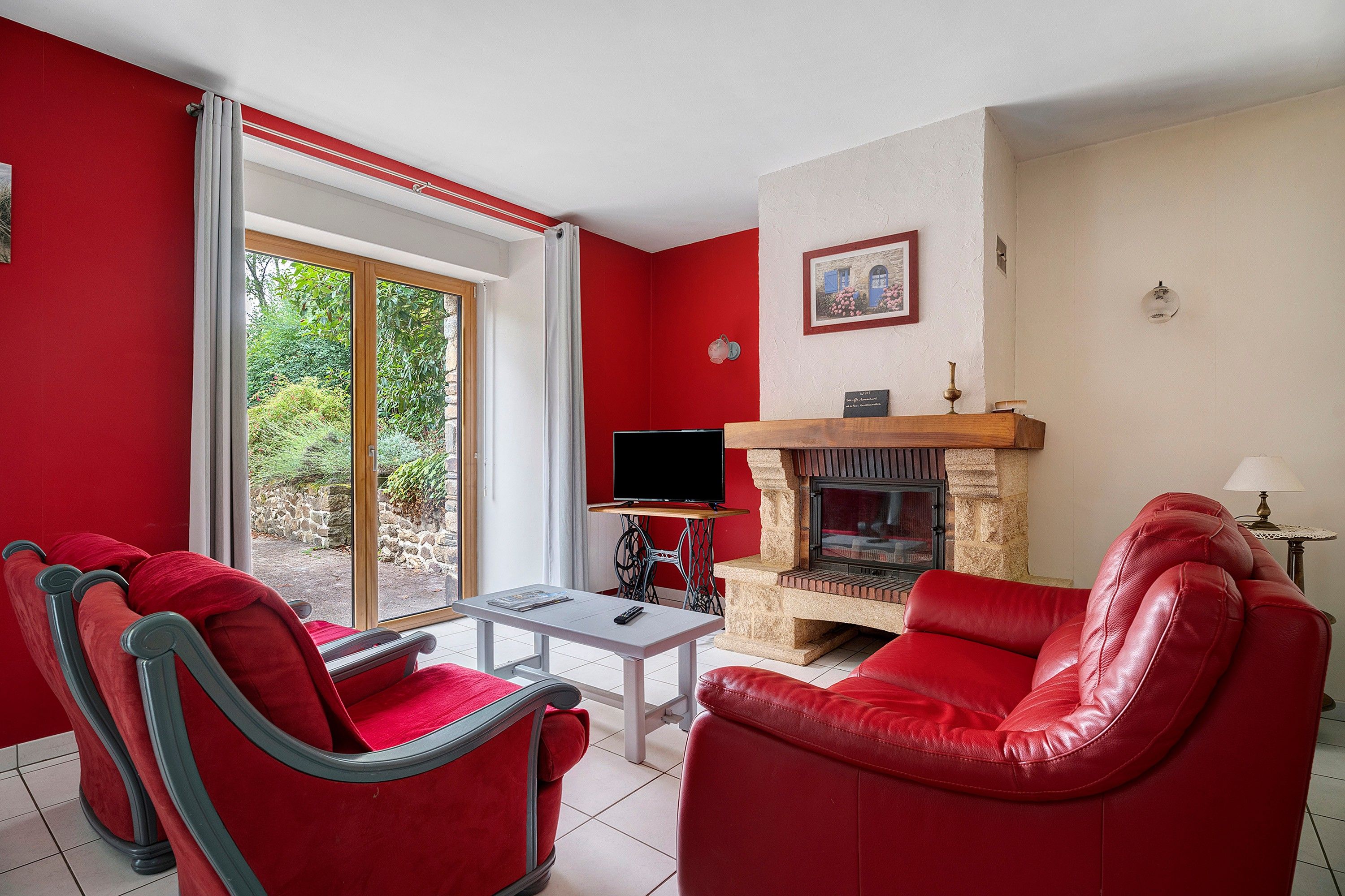 Un salon avec des meubles rouges, une cheminée, un téléviseur et une vue sur la verdure grâce à une porte-fenêtre coulissante.