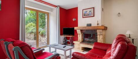 Un salon avec des meubles rouges, une cheminée, un téléviseur et une vue sur la verdure grâce à une porte-fenêtre coulissante.