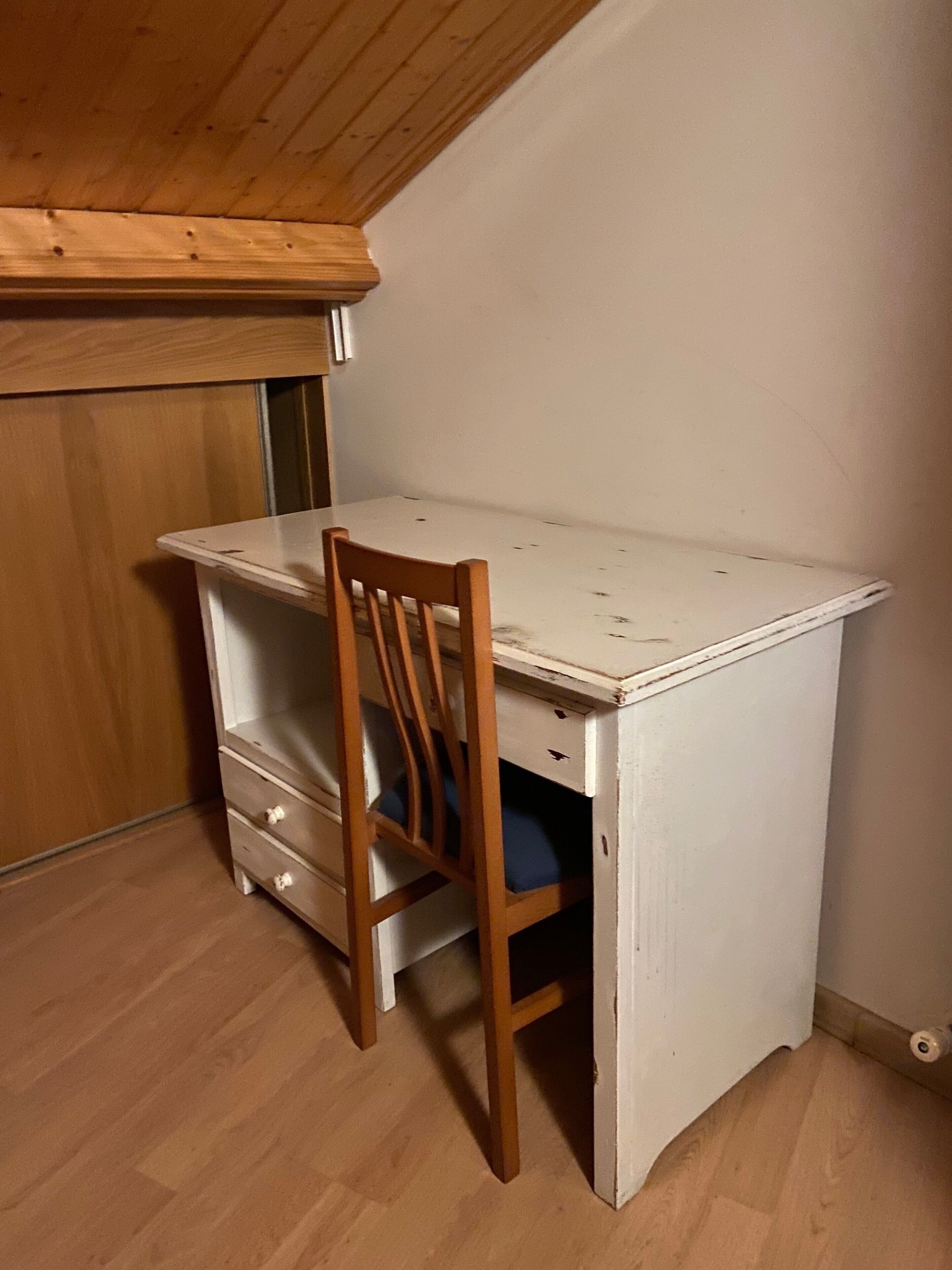 A small, white wooden desk with a chair, a wooden wardrobe, and a wooden floor.
