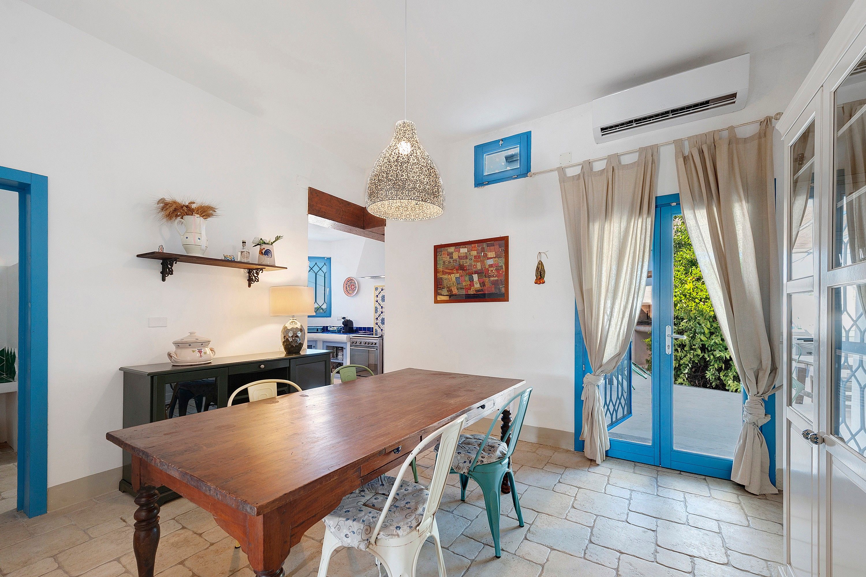 A dining area with a wooden table and chairs, a kitchen in the background, and a view through a blue door.