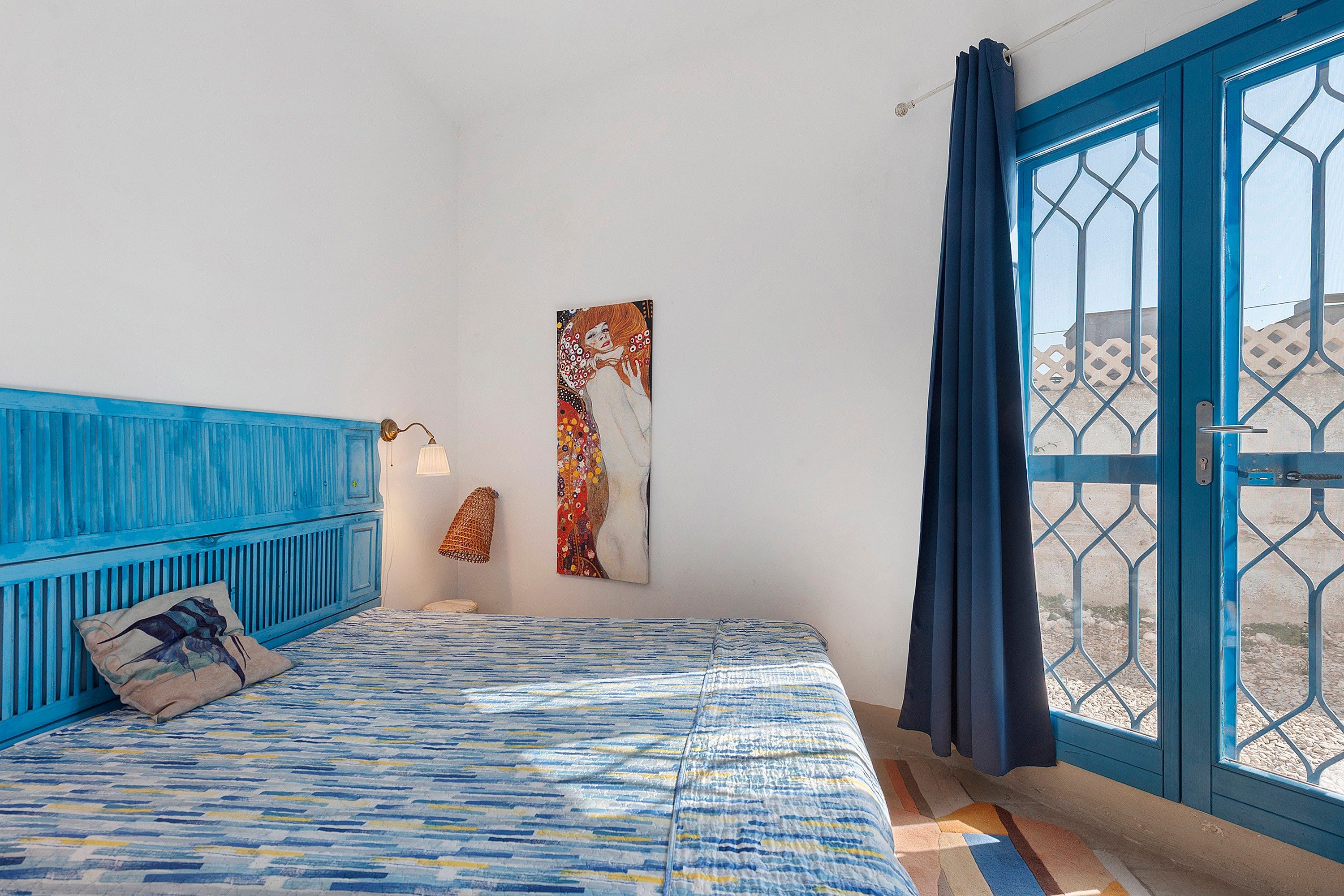 A room with a bed, a wall-mounted lamp, and a large window with a blue door.