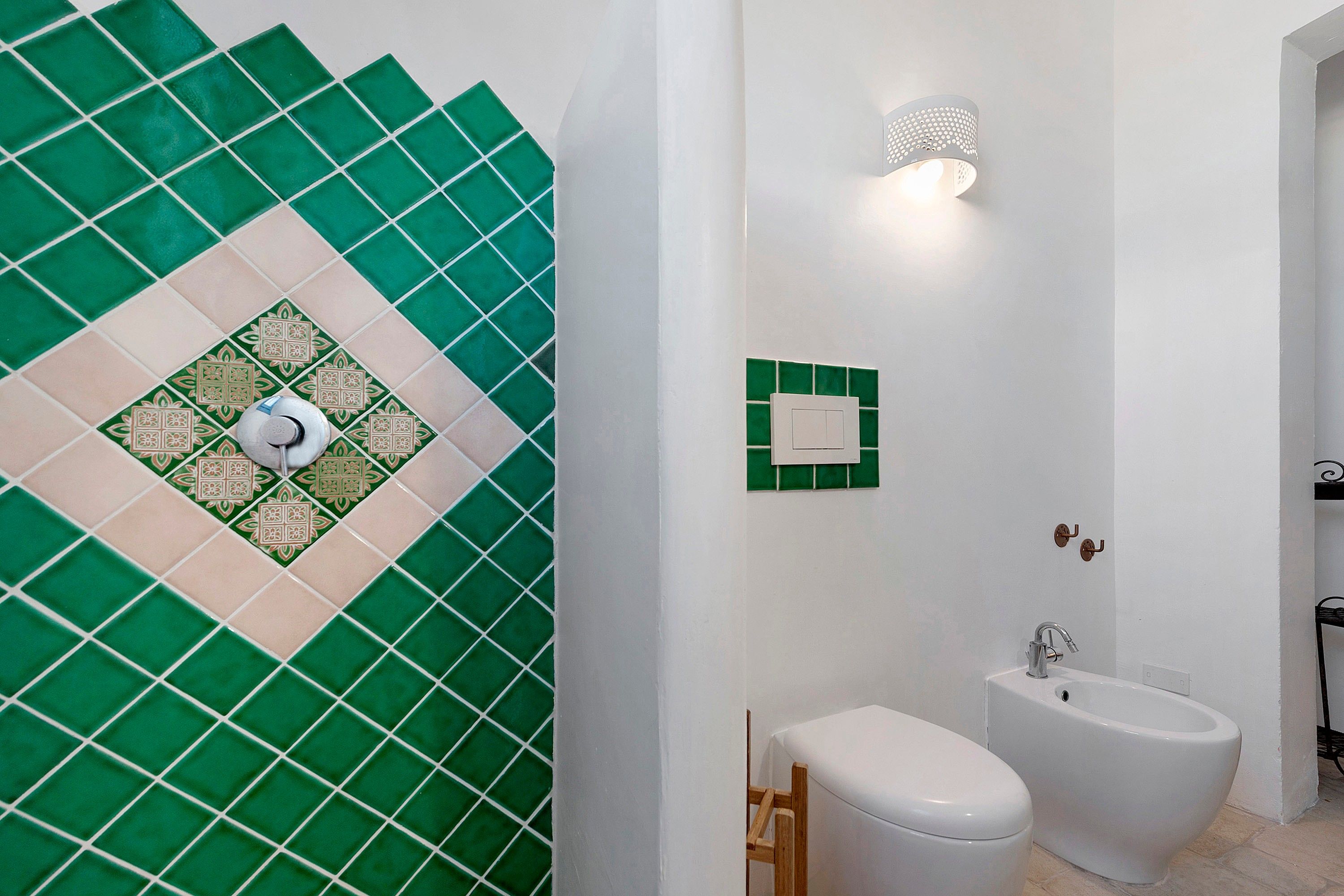 A bathroom with a green and beige tiled wall, a white toilet, a bidet, and a wall-mounted light fixture.