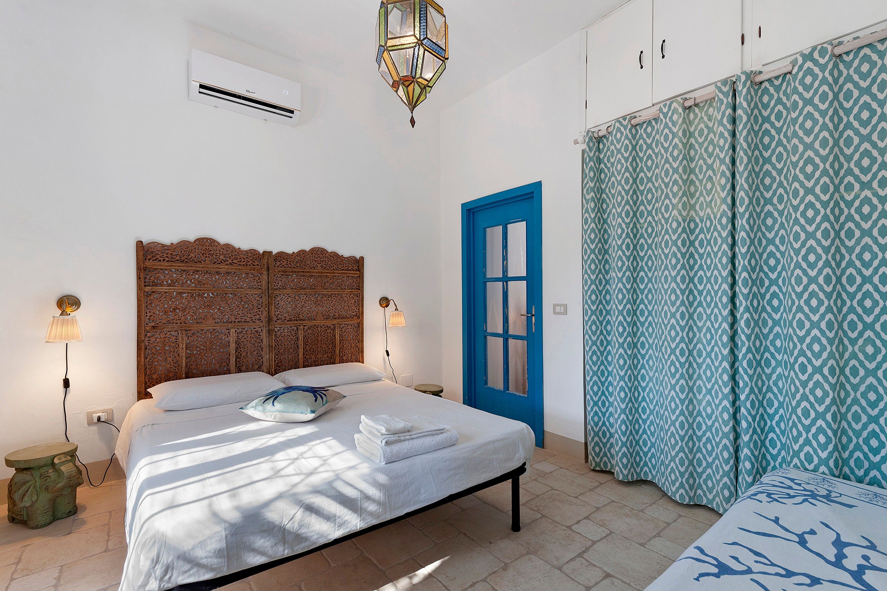 A bedroom with a bed, a blue door, patterned curtains, and a decorative wall panel.