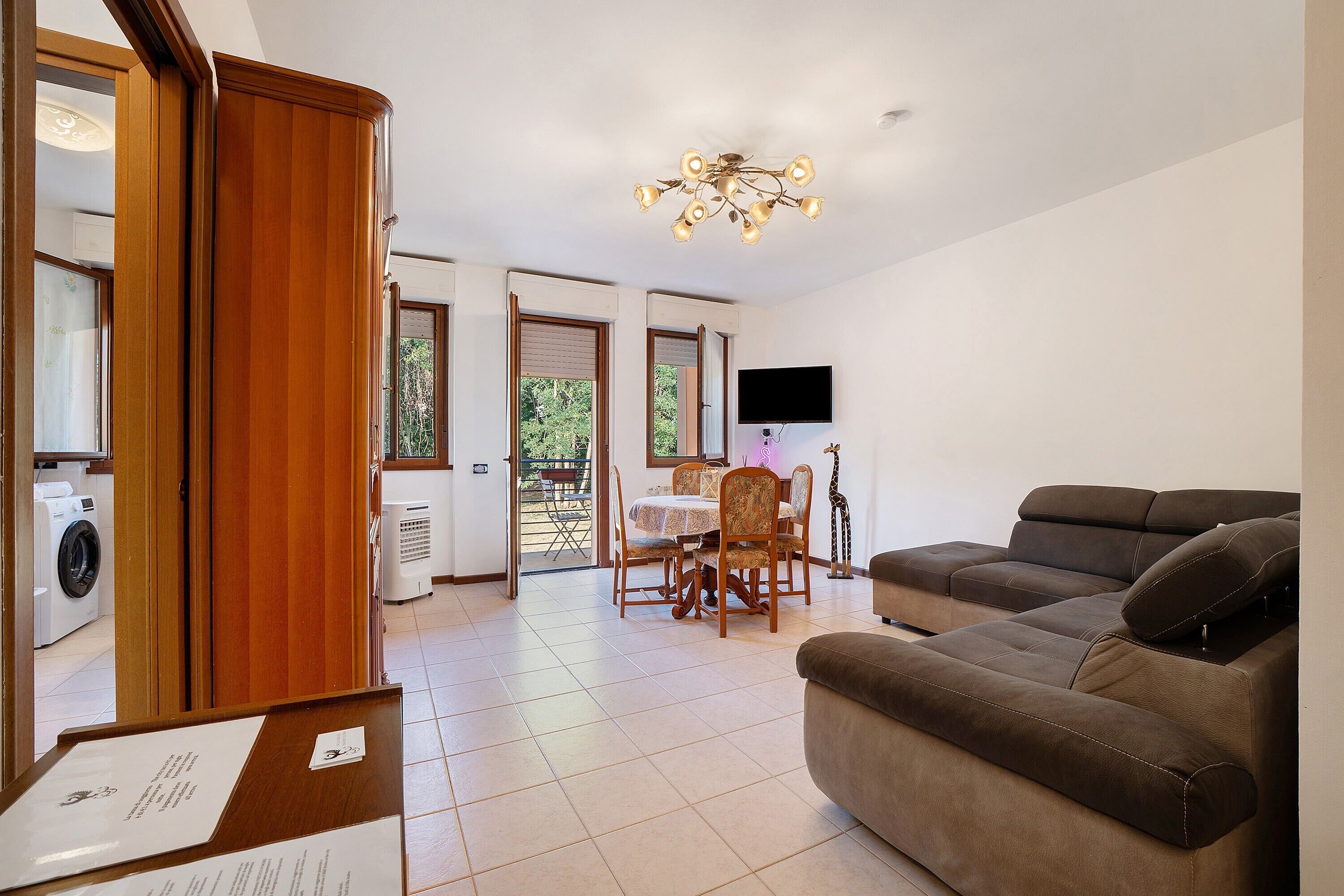 Una sala de estar con un sofá, una mesa de comedor con sillas, un televisor montado en la pared y una araña.