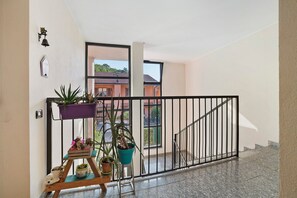 Ein Balkon mit Blumentöpfen, ein Tisch mit Dekoration und Blick auf Gebäude durch ein Fenster.