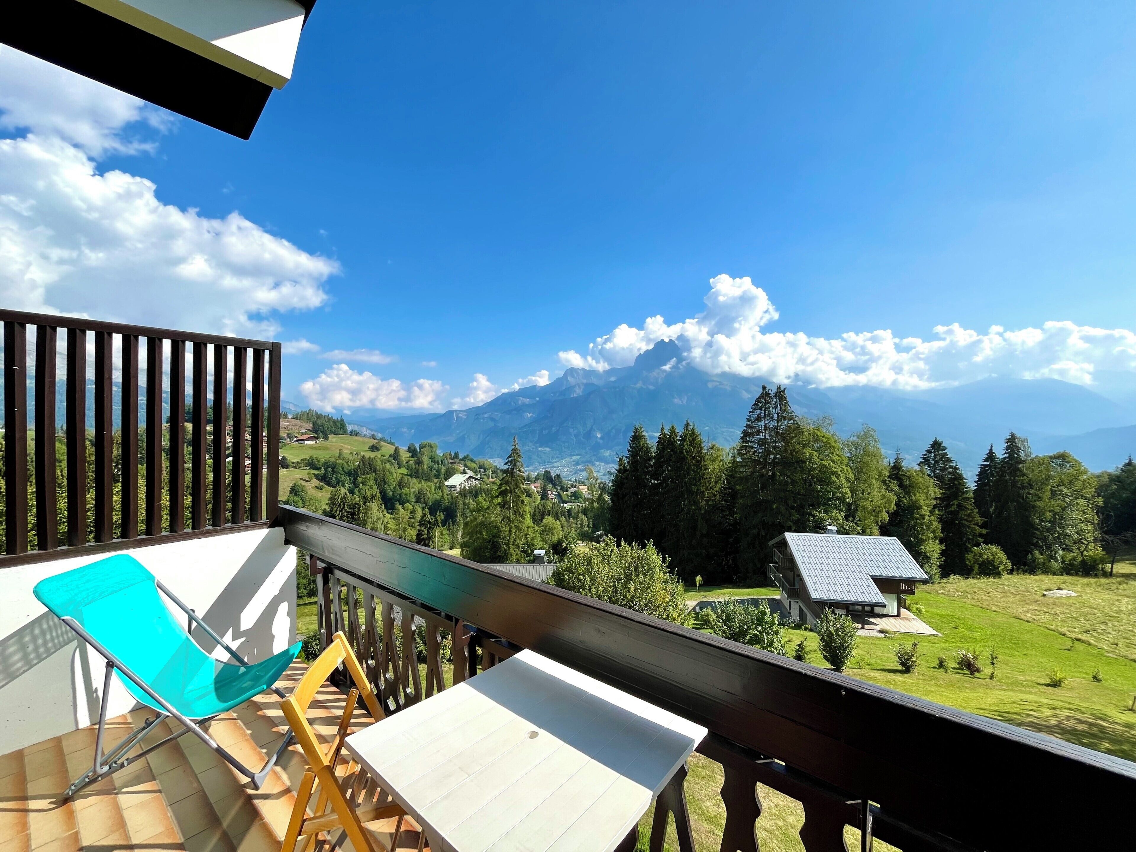 A balcony with a view of a mountain range, a wooden table, and a blue chair.