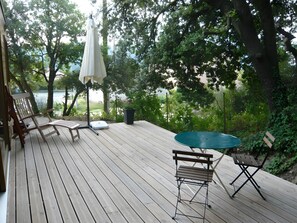 Eine Holzterrasse mit Tisch und Stühlen, einem weißen Sonnenschirm und Blick auf einen von Bäumen umgebenen See.