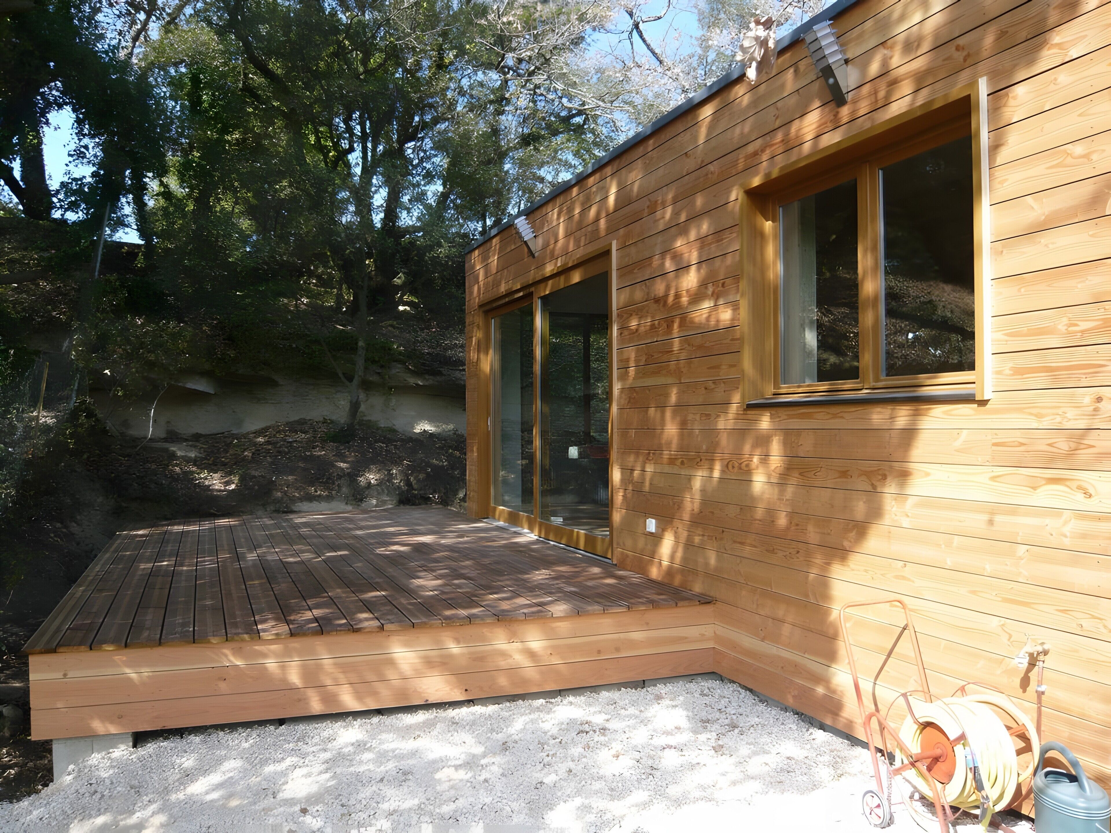 Une cabane en bois dotée d’une terrasse, de grandes fenêtres et d’un enrouleur de tuyau d’arrosage.