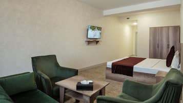 A hotel room with a green sofa, a wooden table, a bed with a brown blanket, a television on the wall, and a wardrobe.