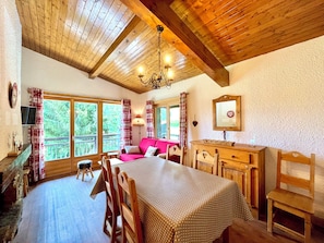 Ein gemütliches Zimmer mit Holzbalkendecke, einem Esstisch mit Stühlen, einer Couch und einem Fenster mit Vorhängen.
