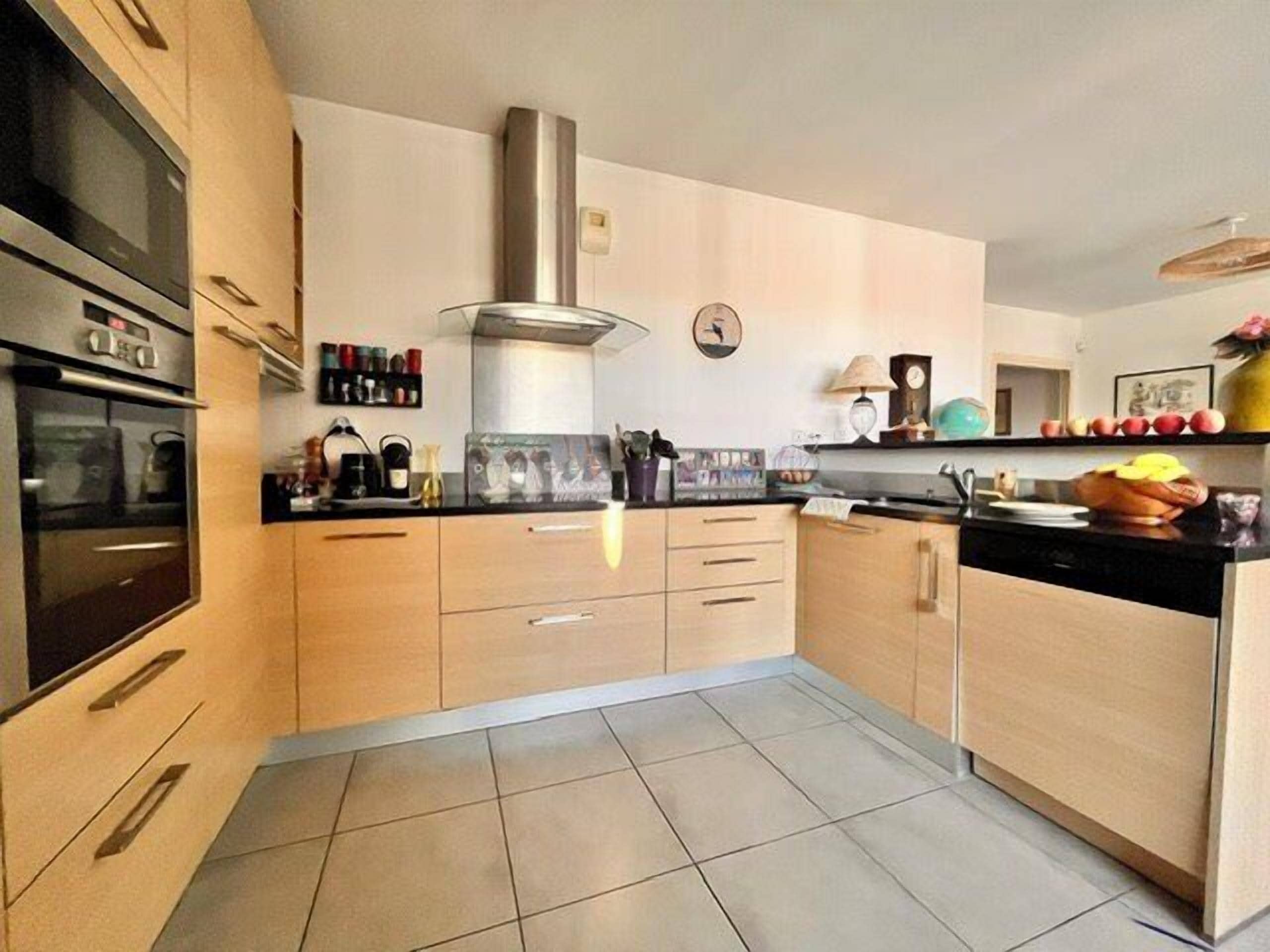 A modern kitchen with light wood cabinets, a stainless steel range hood, and a tiled floor.