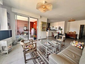 Ein Wohnzimmer mit Sofa, Couchtisch und Sesseln, einem Fernseher und einer Glastür, die auf einen Balkon führt.