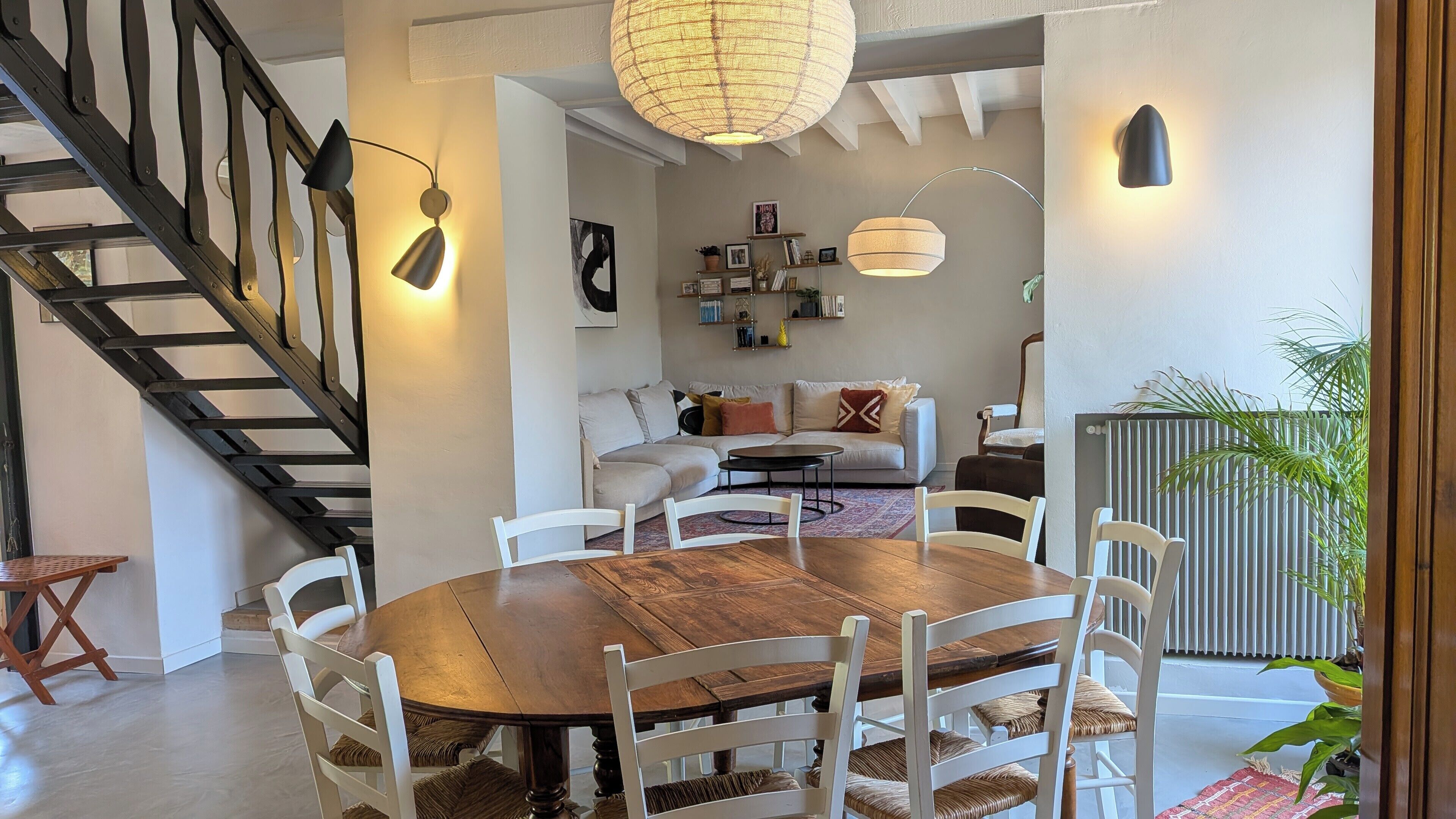 Una zona de menjador amb una taula de fusta i cadires blanques, una escala amb baranes negres, i un saló amb un sofà i objectes decoratius.