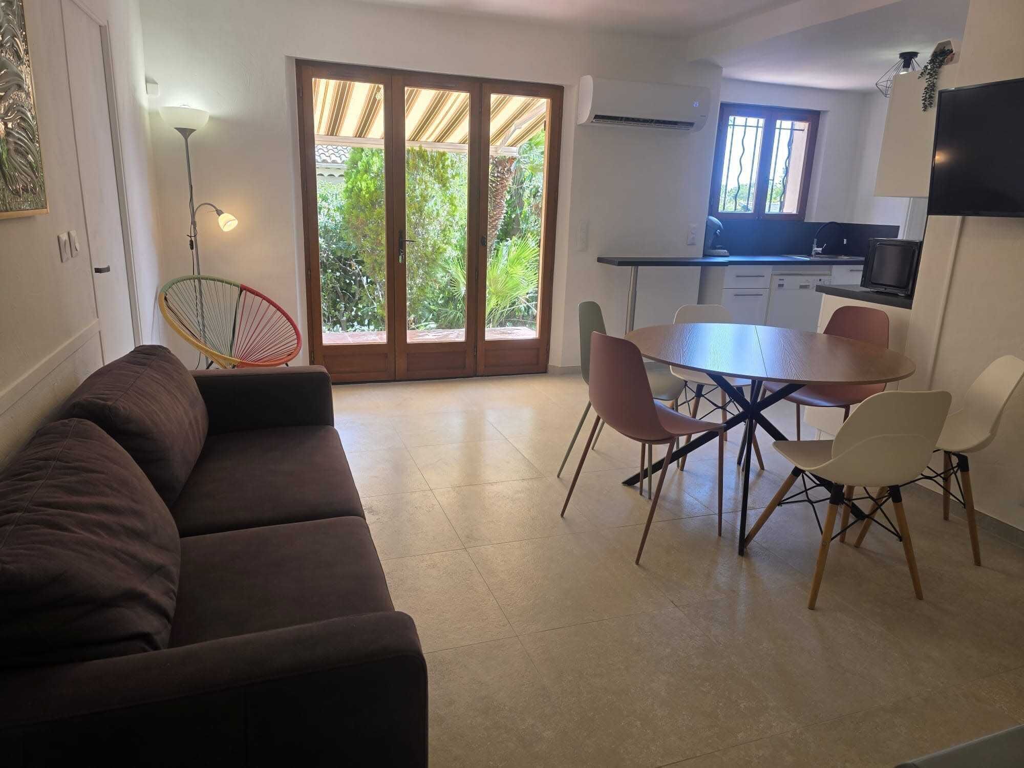 A modern living room with a sofa, dining table, and chairs, a kitchen with a TV, and a sliding glass door to a garden.