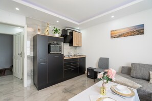 Une cuisine moderne équipée d’un four, d’un micro-ondes et d’une cuisinière intégrés, un coin repas avec une table dressée pour deux personnes, et un salon avec un canapé et un tableau au mur.
