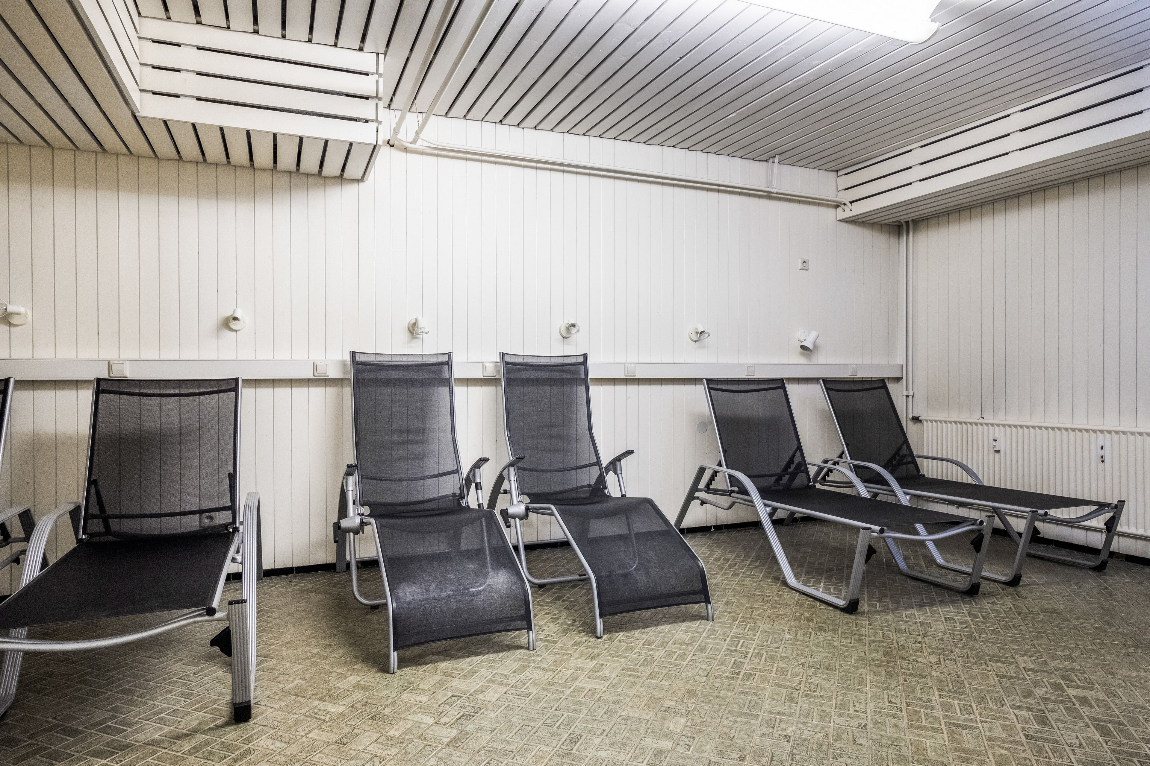 A waiting area with four reclining chairs, a ceiling with ventilation, and a white wall with a radiator.