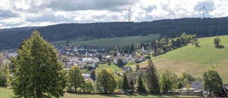 A town with houses, a forested hill, and wind turbines in the background.