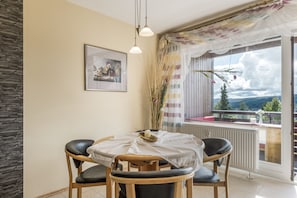 A dining area with a table and chairs, a framed picture on the wall, and a view of the outdoors through a window. - Apartment 'Beautiful View with Pool', Balcony & Wi-Fi (Schonach im Schwarzwald)
