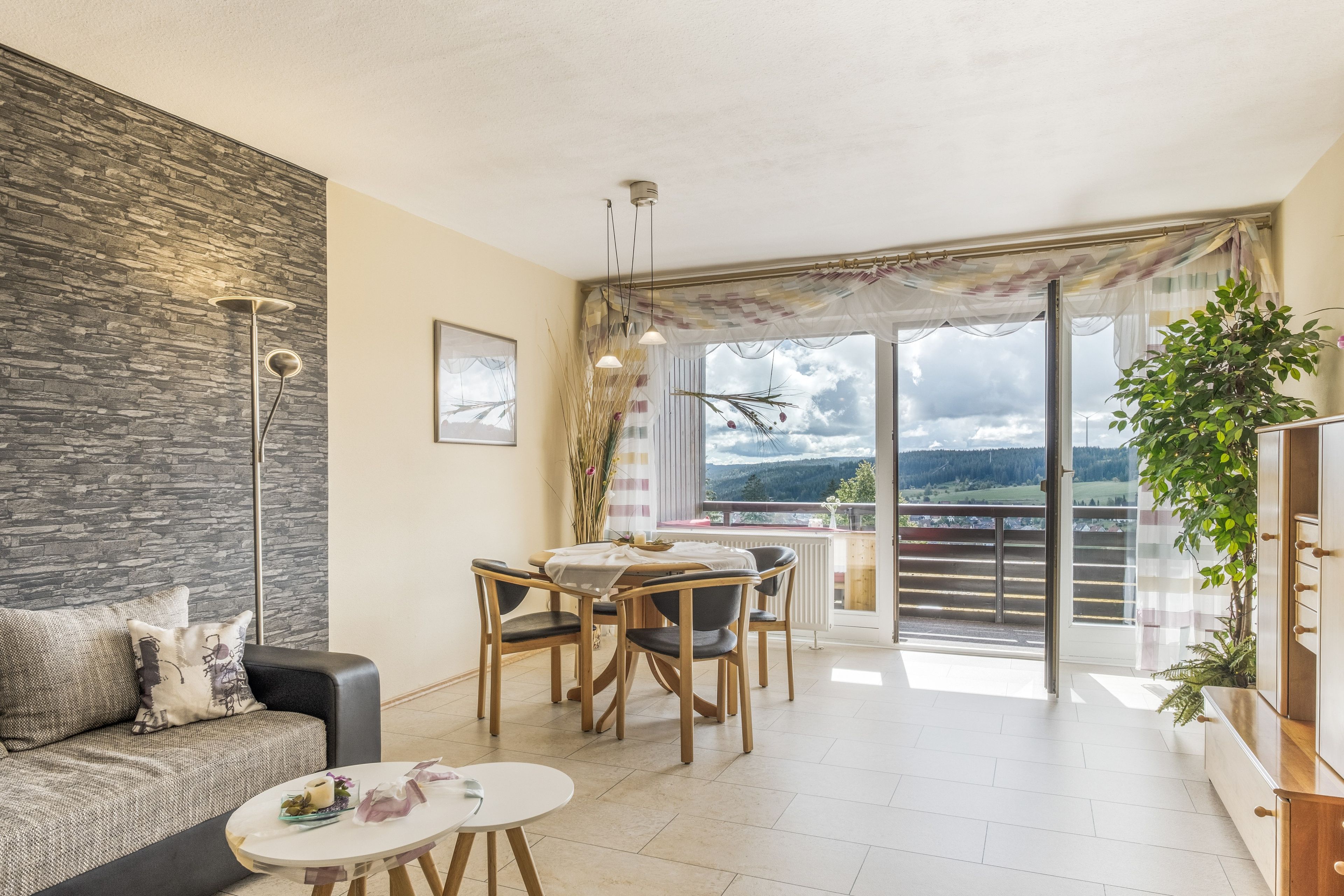 Una sala de estar moderna con una pared de piedra, una zona de comedor con mesa y sillas, y un balcón con vistas al campo.