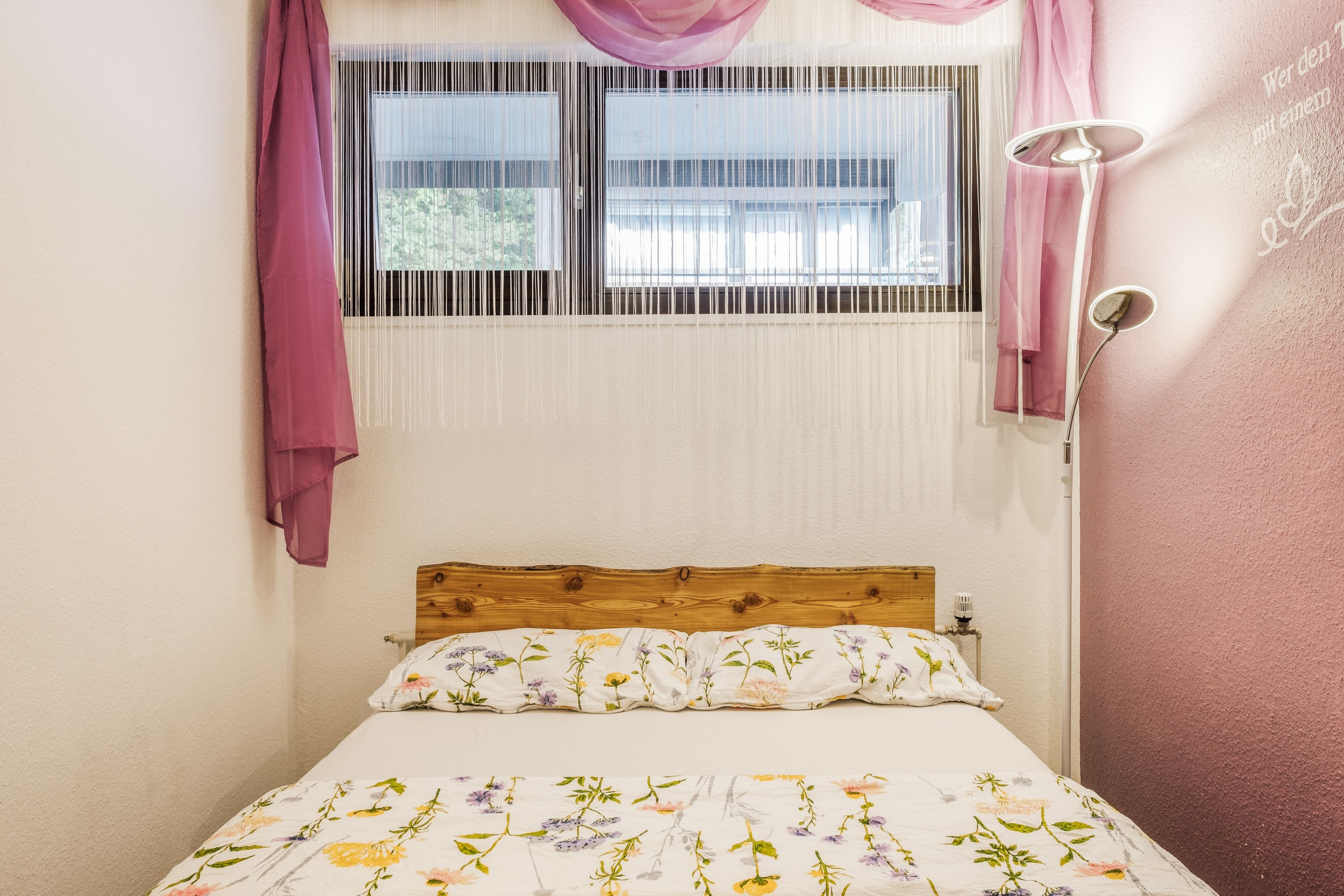 Une chambre à coucher avec une tête de lit en bois, une couvre-lit à motifs floraux et une fenêtre équipée de stores verticaux.
