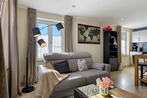 Un moderno salón con un sofá gris, una mesa de centro con un jarrón de flores, un mapa del mundo en la pared y una cocina al fondo.