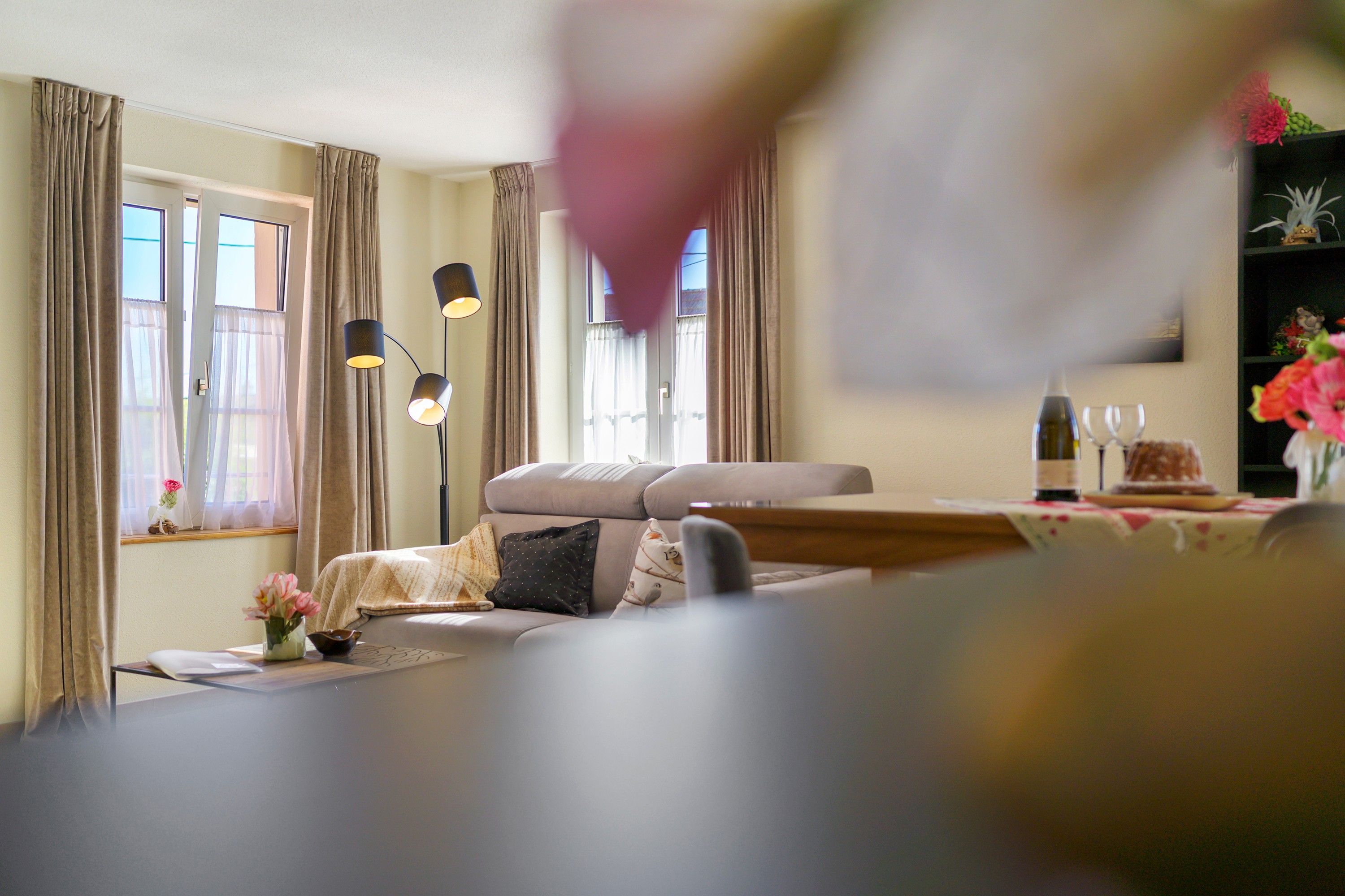 Een woonkamer met een bank, een bijzettafeltje en een boekenkast. Er hangen gordijnen voor de ramen, en op het tafeltje staat een lamp.