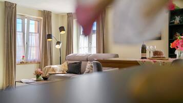 Een woonkamer met een bank, een bijzettafeltje en een boekenkast. Er hangen gordijnen voor de ramen, en op het tafeltje staat een lamp.