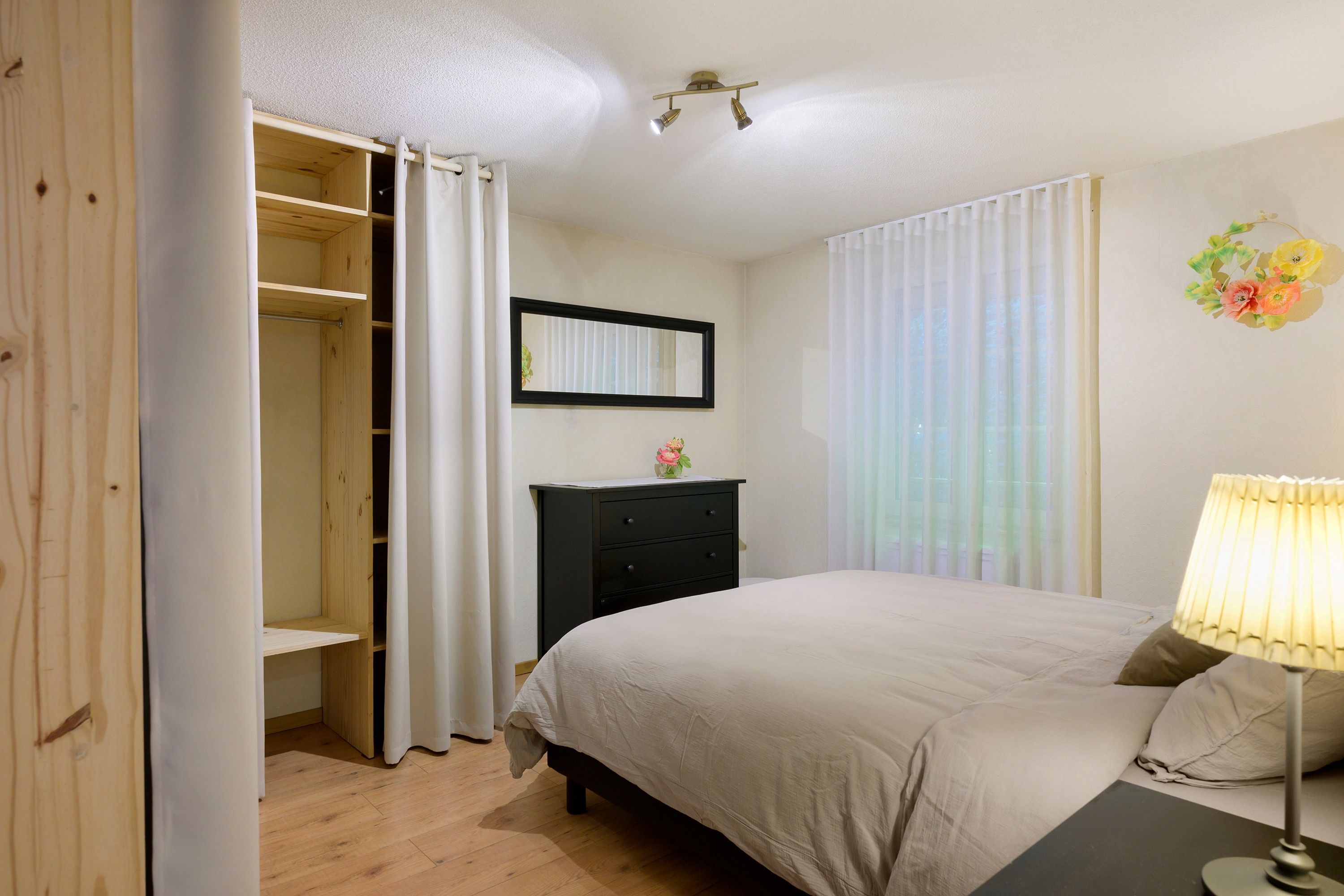Une chambre à coucher avec un lit, une commode, un miroir et une fenêtre avec des rideaux.