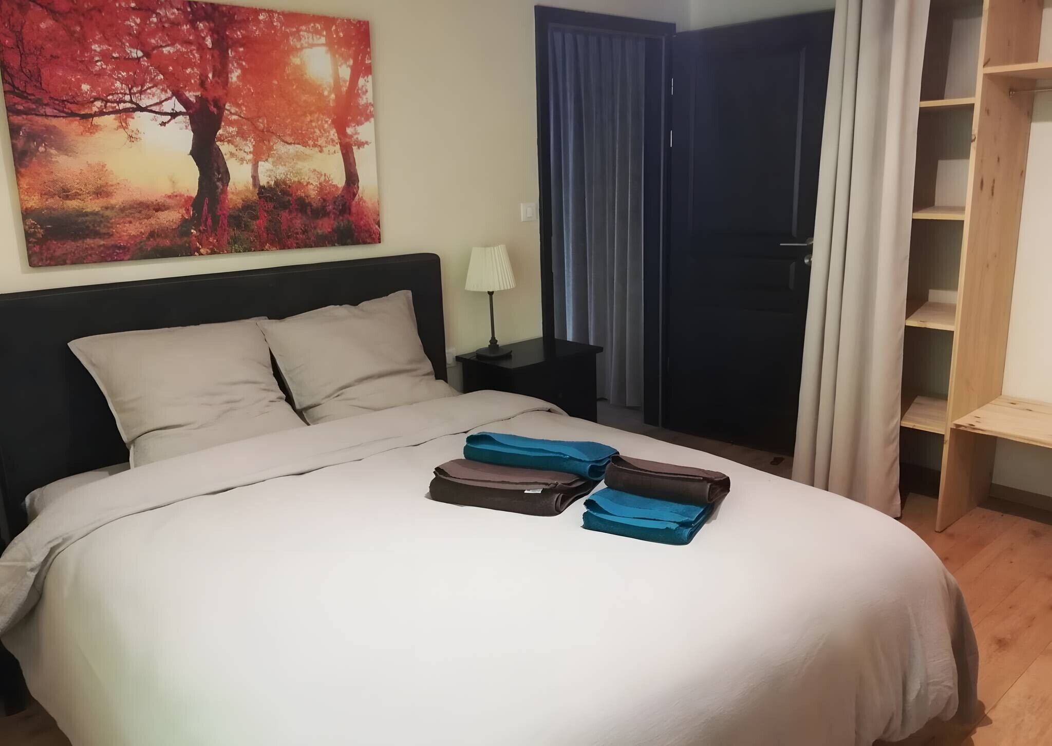 Een netjes opgemaakt bed met kussens en een opgevouwen plaid, een bedlamp, een schilderij van bomen, en uitzicht op een balkon met gordijnen.
