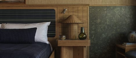 A bedroom with a wooden headboard, a bedside table with a lamp, a rug, and a small shelf with books.